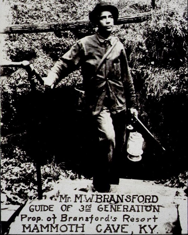 A black and white photo of a man standing near the entrance of the cave.