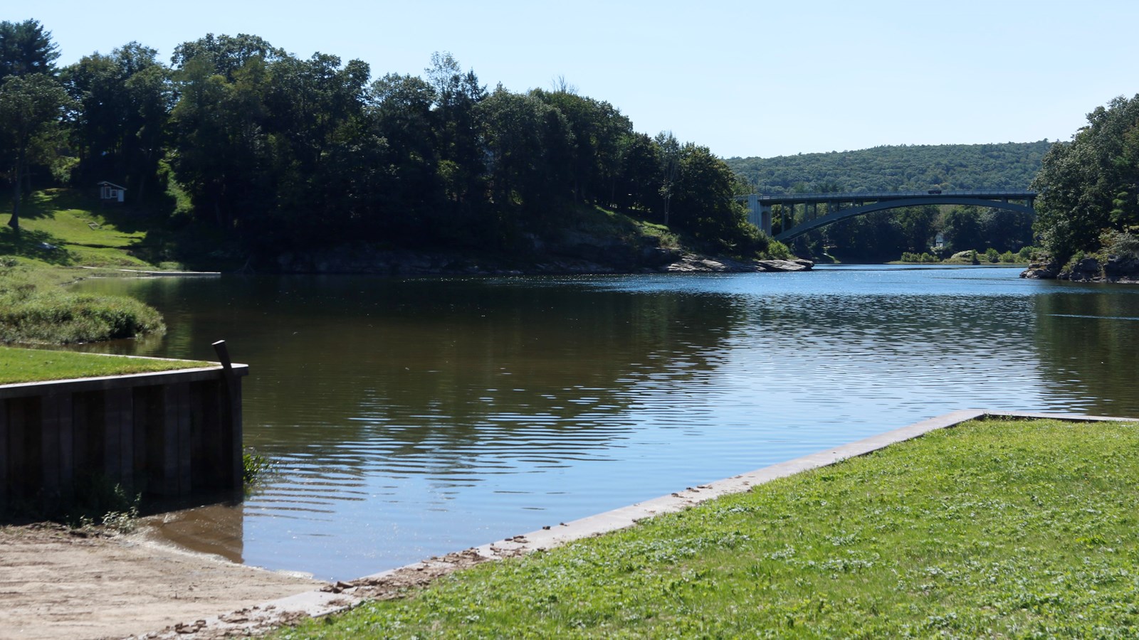 View of river with bridge between 2 hills in distance. In foreground, concrete ramp leads to water.