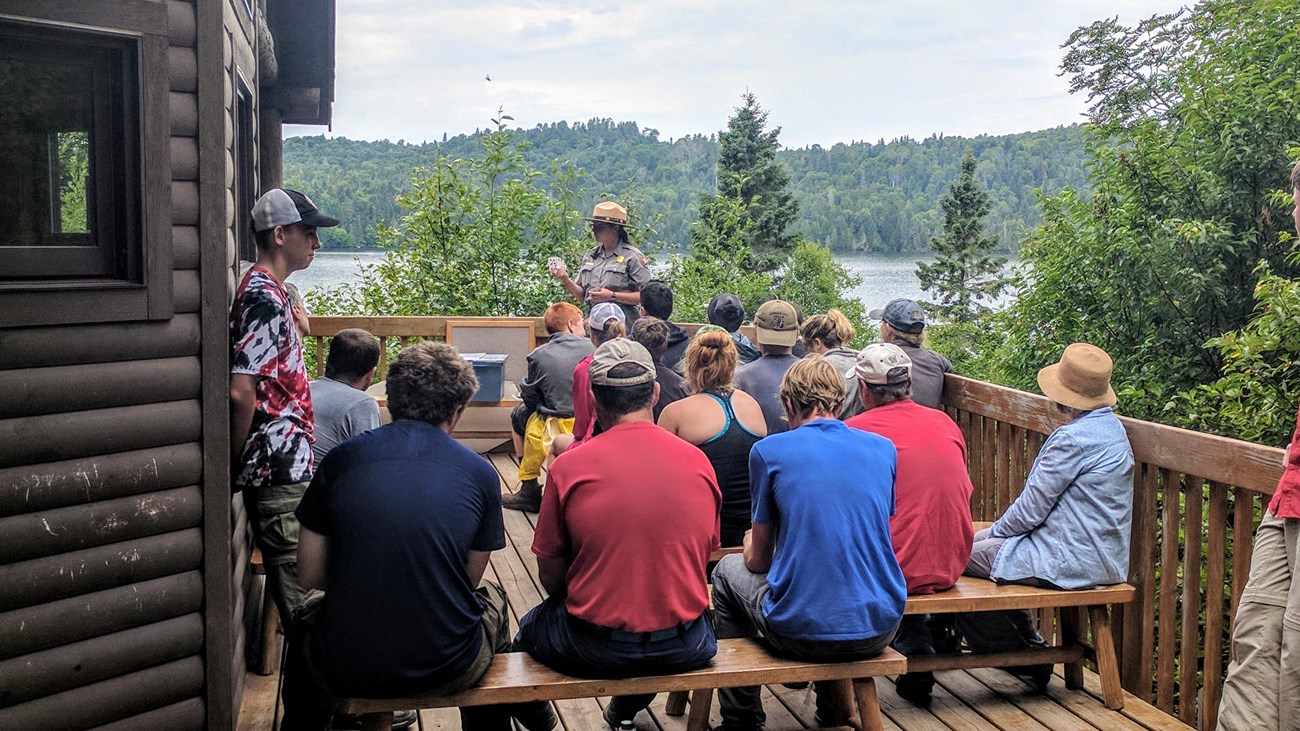 A park ranger gives a program to a group of people on a porch with Washington Harbor behind.