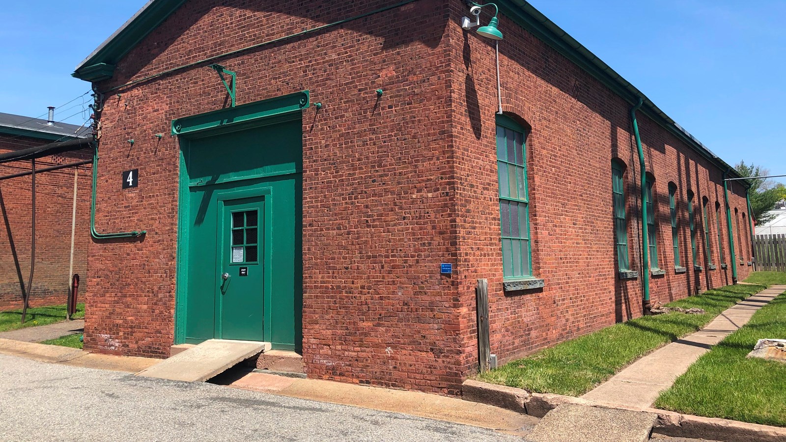 Long, rectangular, one story, red brick building, with green door. Grass and sky surround it.