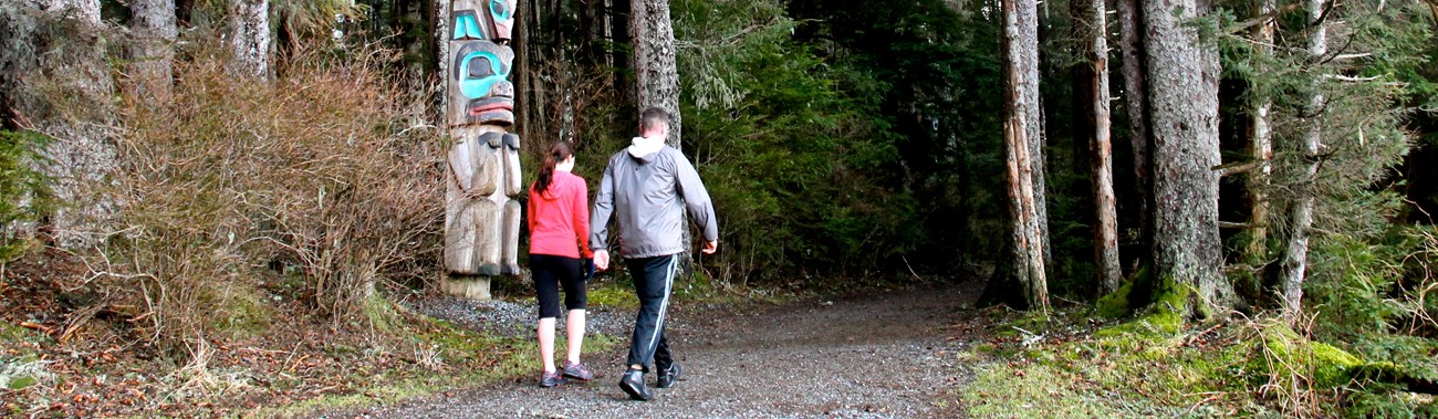 A man and woman walk by trees and a totem pole on a gravel path