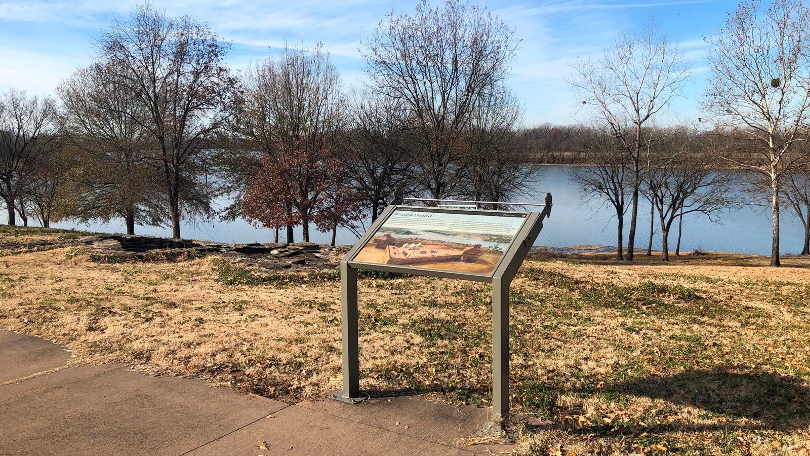 Wayside panel next to a sidewalk. The Arkansas River and leafless trees in the background.