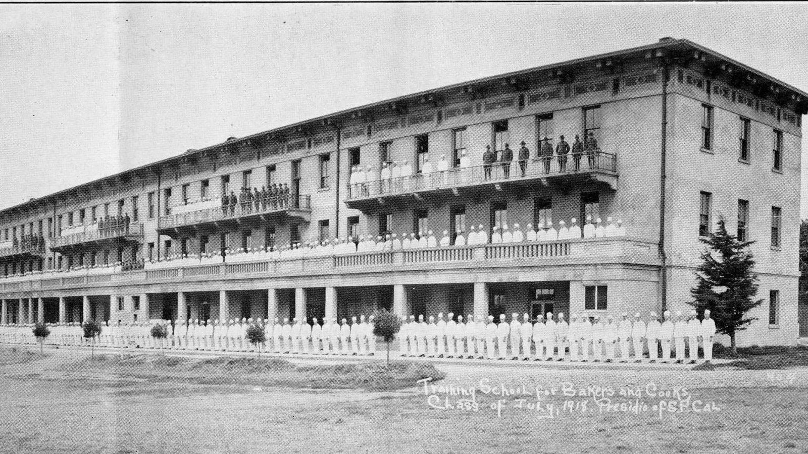 Men in white stand in front of and on the balconies of a three story building.