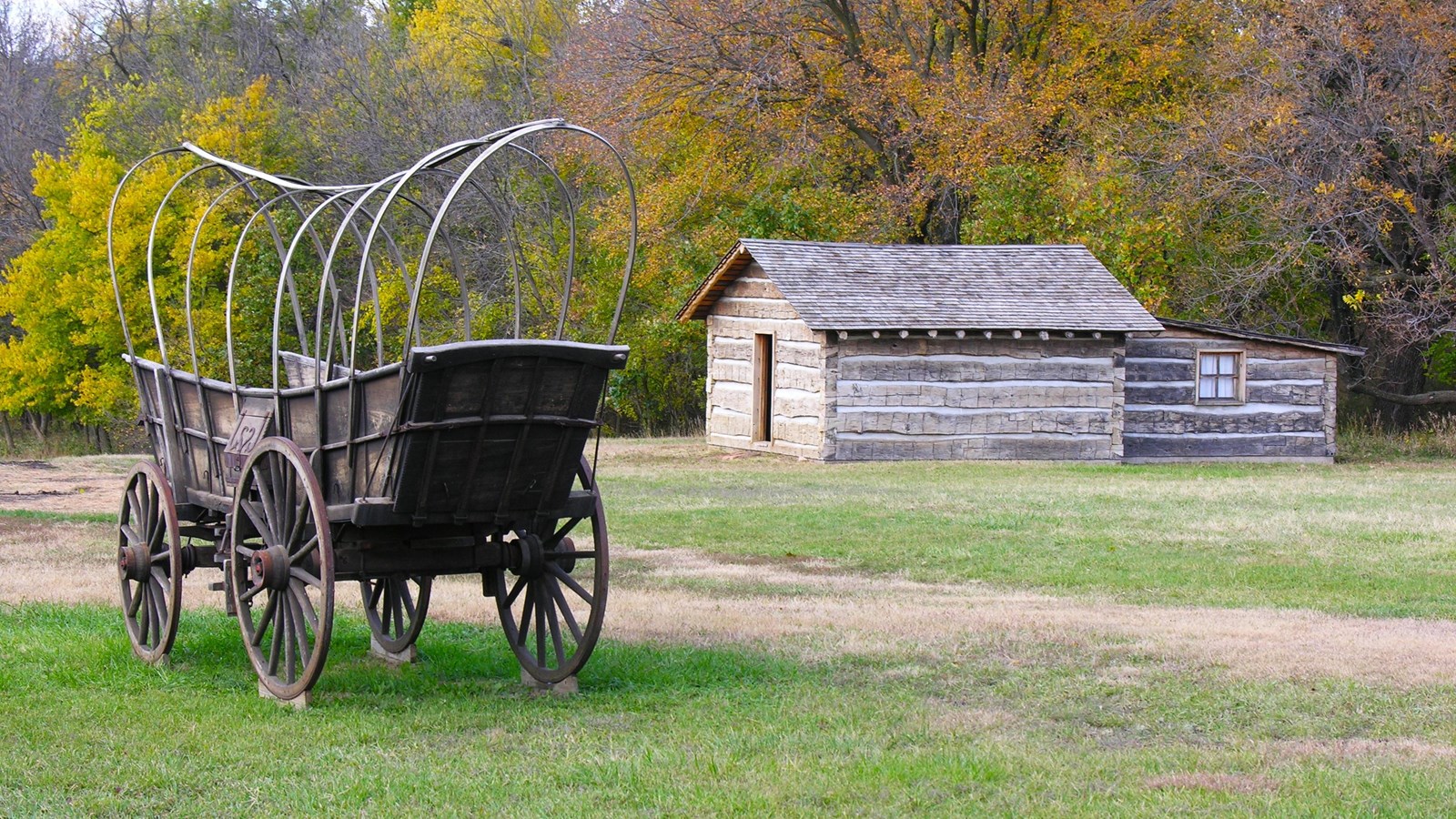 An unpainted wagon with red hubs and bare hoops. Nearby to right stands log building with door.
