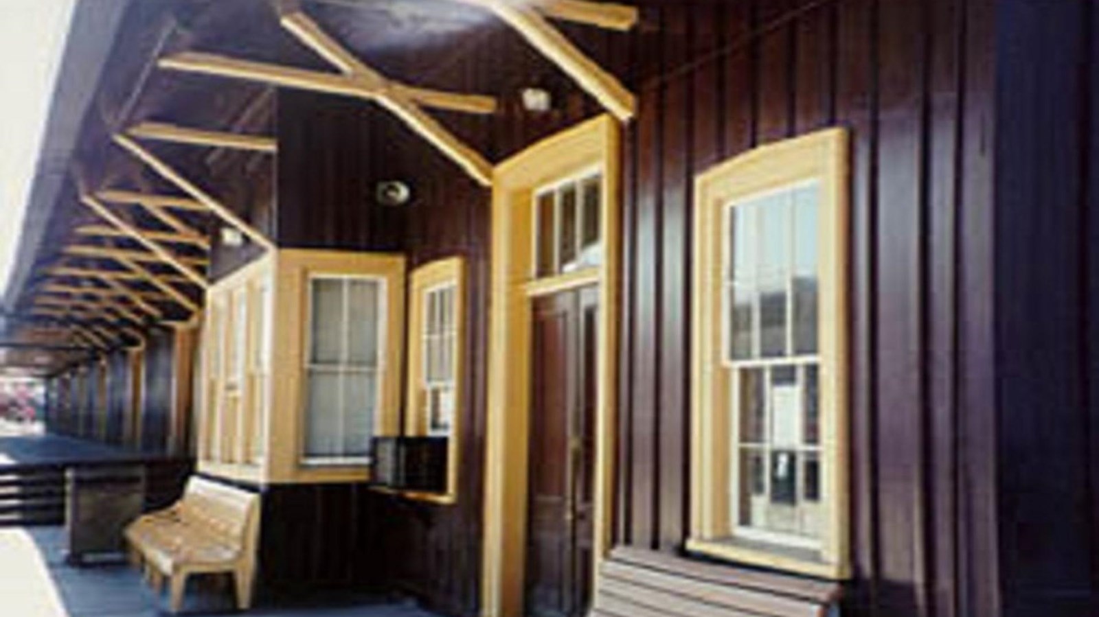 Exterior of a wooden building with dark brown panels and yellow-trimmed windows, featuring a bench a