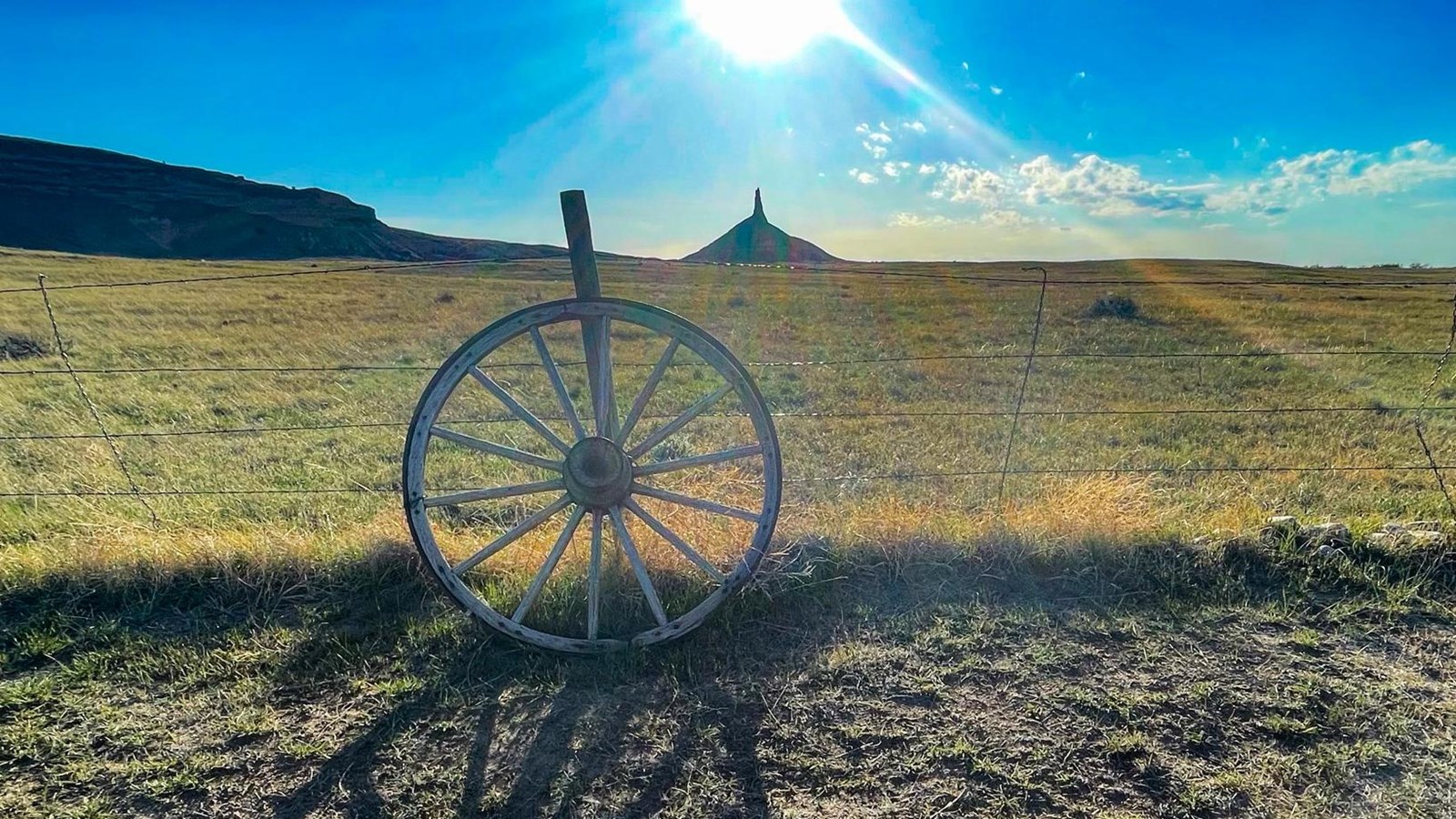 A wagon wheel rests on barbed wire fence with Chimney Rock and blazing sun in the background.