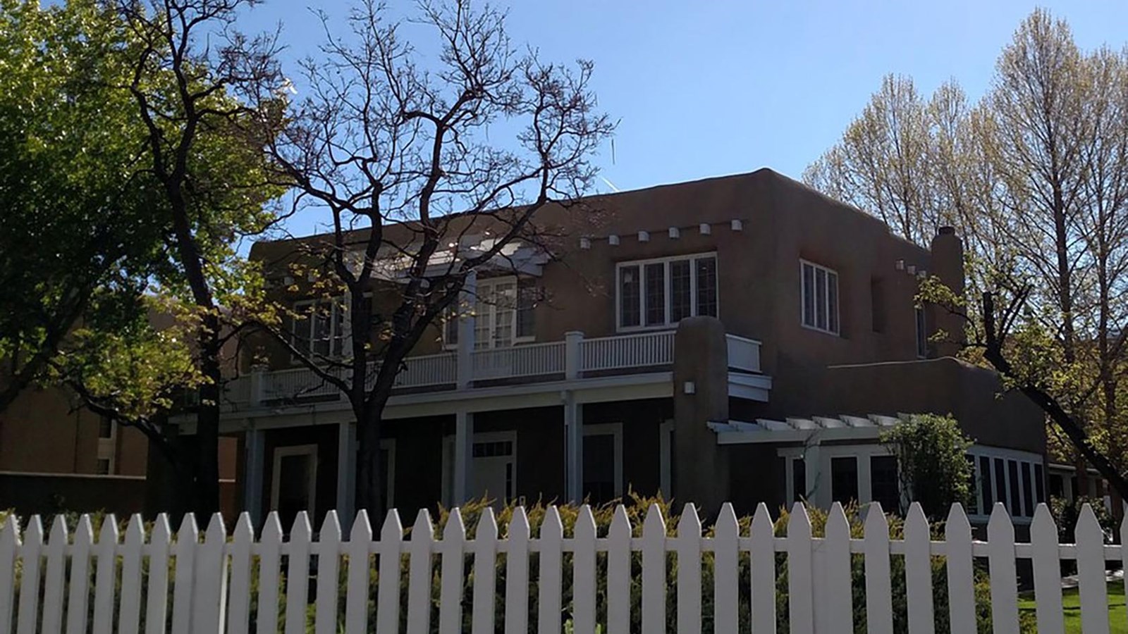 White picket fence in front of a two story adobe house with a white upstairs veranda.