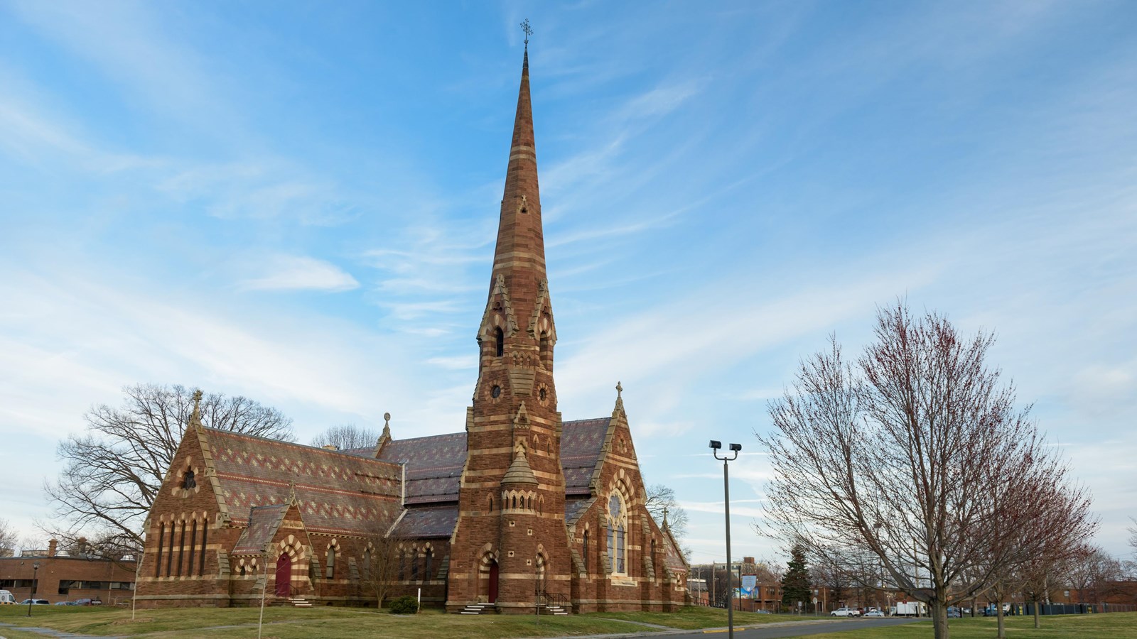 A Gothic Revival Church with a spire on a cloudy day with a green lawn and bare trees.