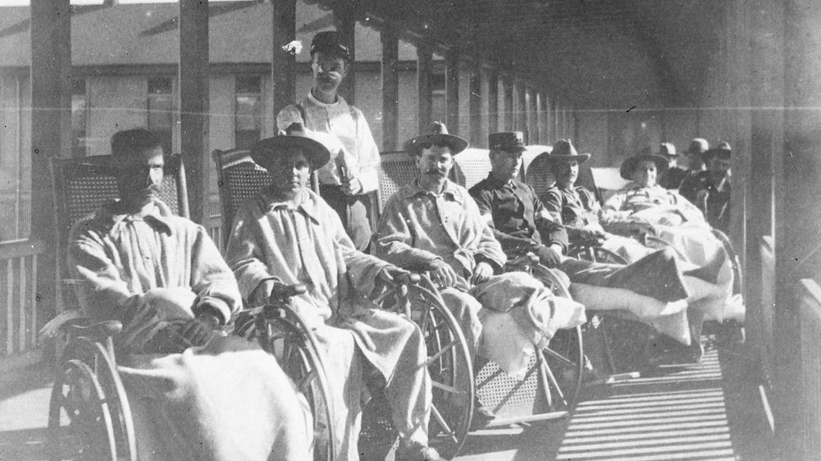 Black and white photograph of several men in wheelchairs sit on a porch. 