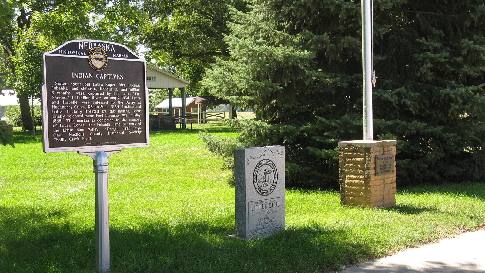 A brown sign on a silver post, headed “Indian Captives,” stands by a granite Pony Express monument.