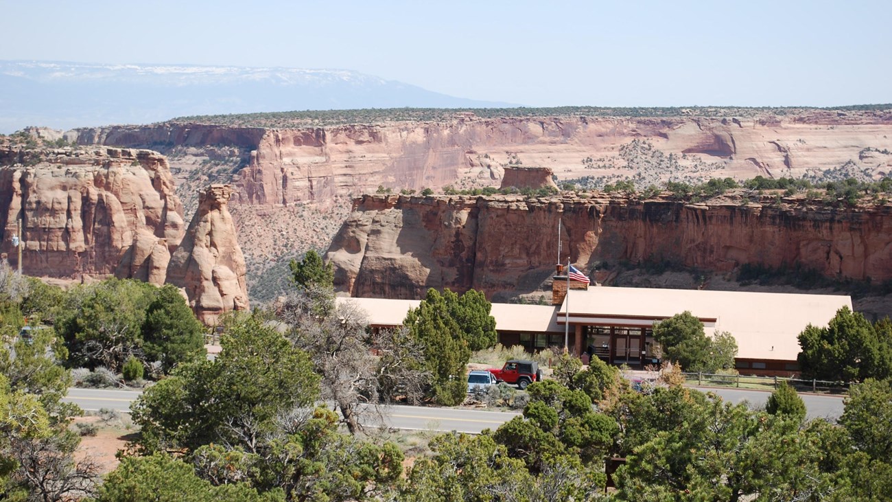 A low, mostly flat building with a flagpole and shrubs, standing before a desert canyon