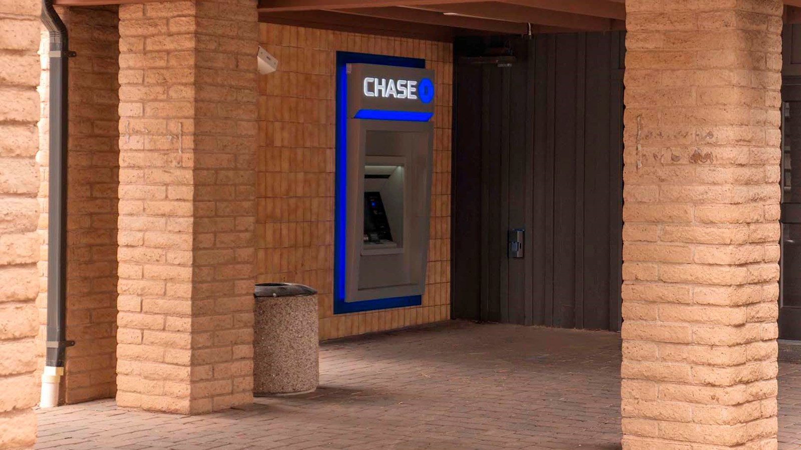 Looking between beige brick pillars down a breezeway at an ATM machine with the word,