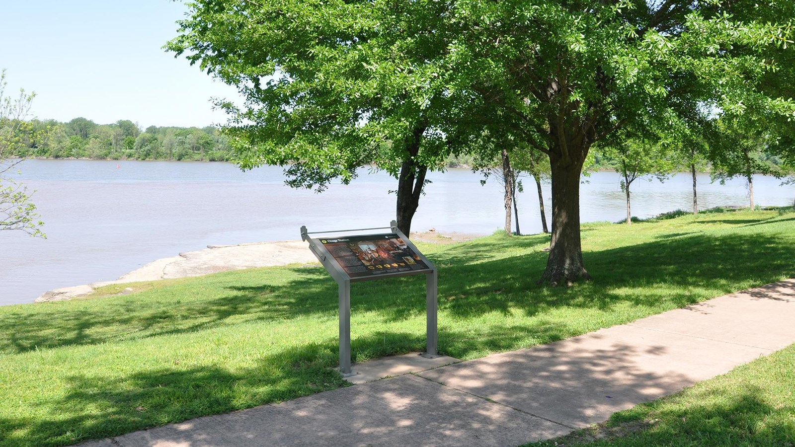 Wayside on edge of sidewalk with grass sloping gently toward the rocky banks of the Arkansas River.