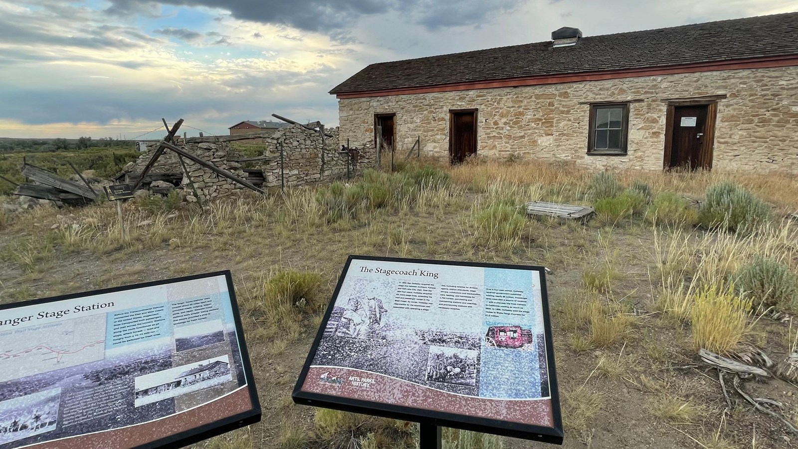 Two exhibit panels sit in front of a one story, stone building.