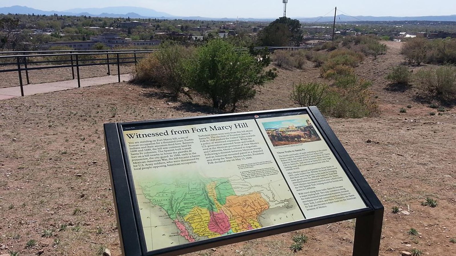 A wayside exhibit looking out towards a ruin of an adobe wall, on a bluff overlooking a city.
