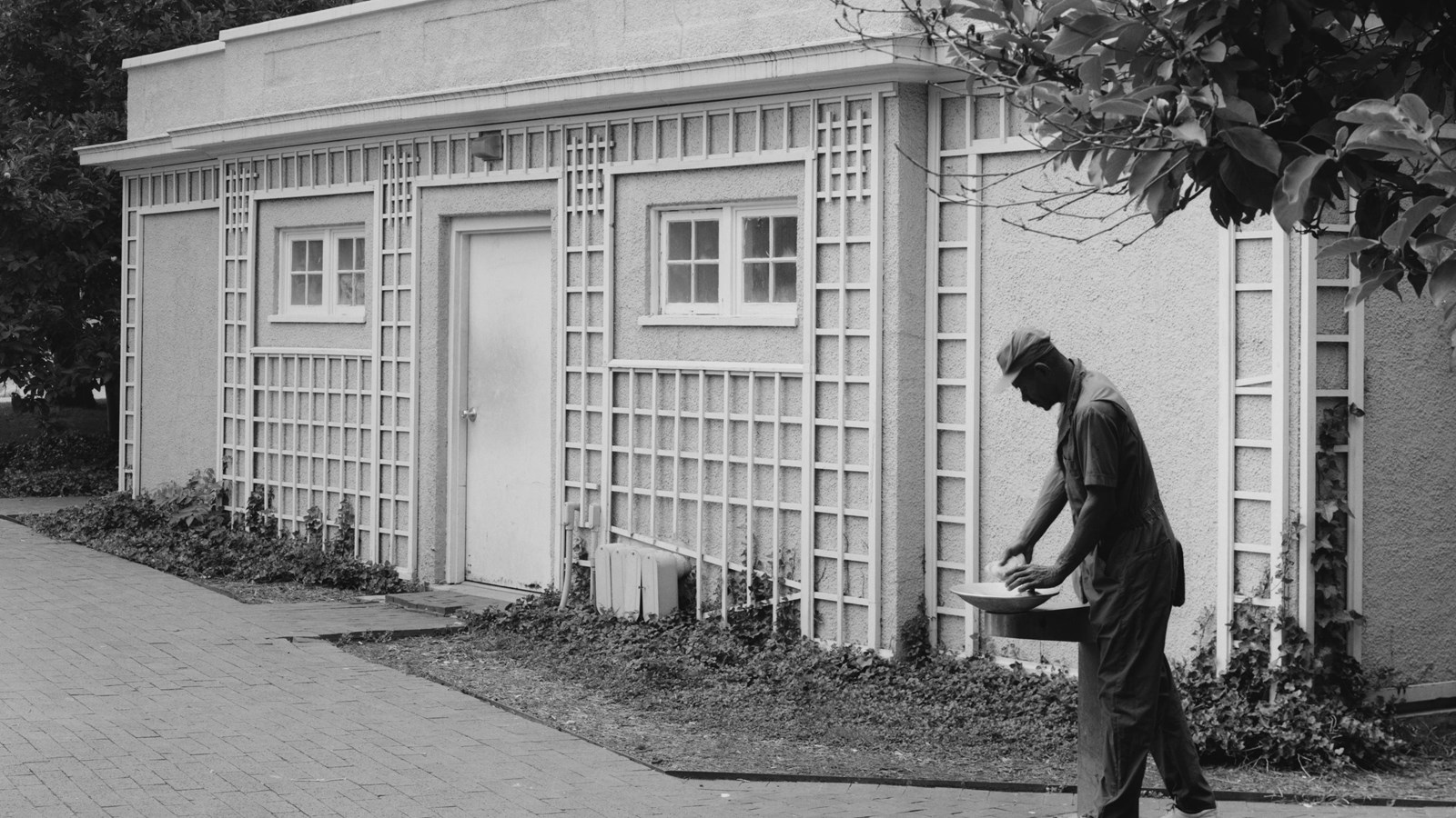 A man fills up his water bottle at a water fountain in front of a squat one-story latticed building.