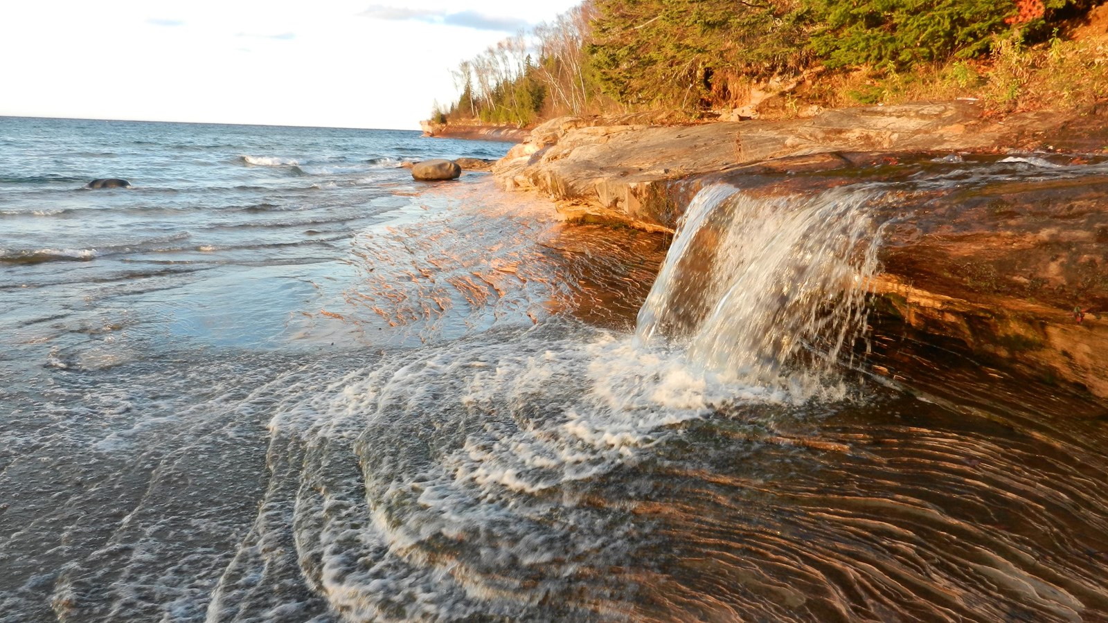 Two foot tall waterfall flows over a rocky ledge and slope of smooth rock into Lake Superior.