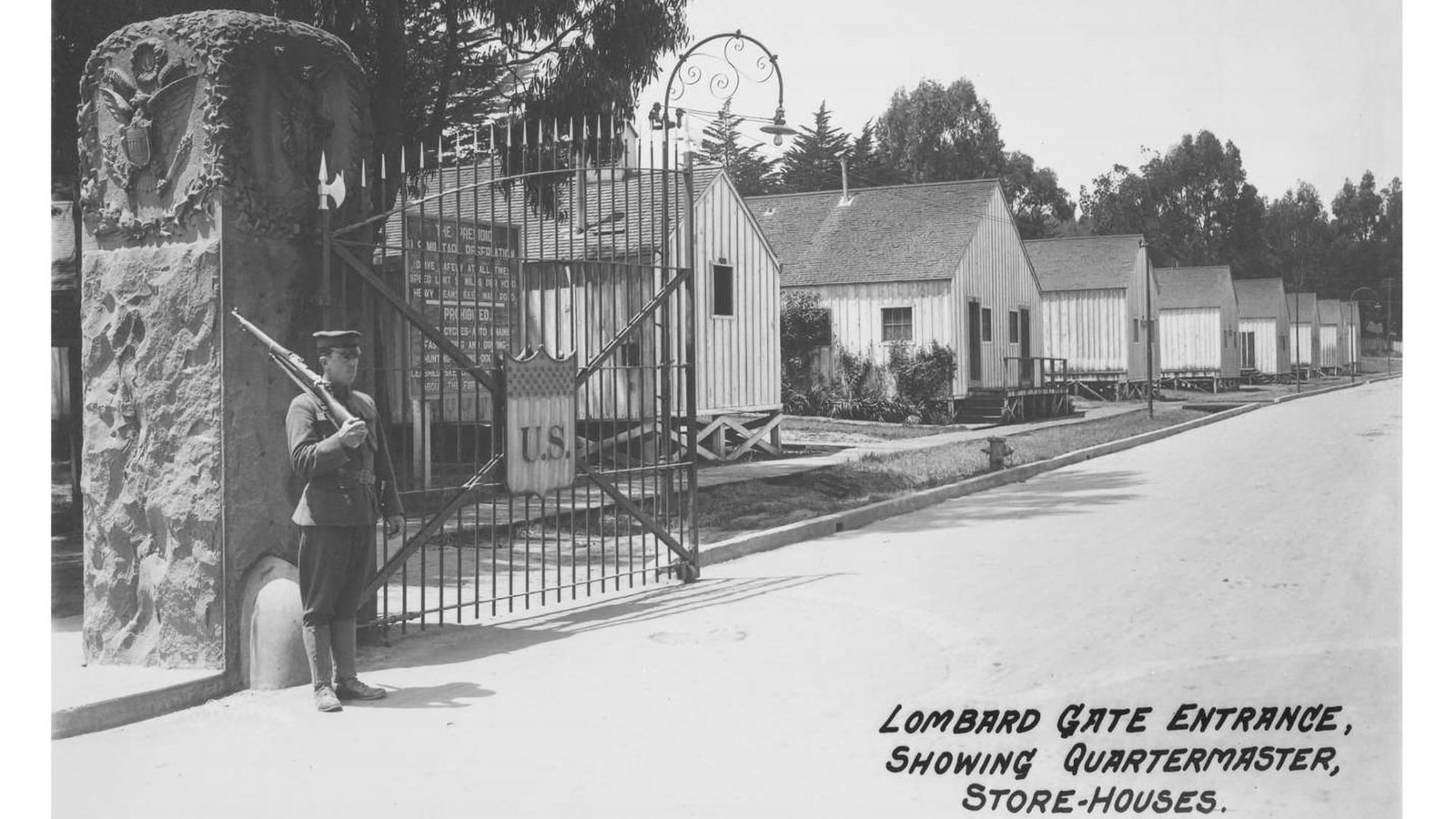  A man with a gun stands  in front of a gate. Rows of wooden buildings line the street behind him.