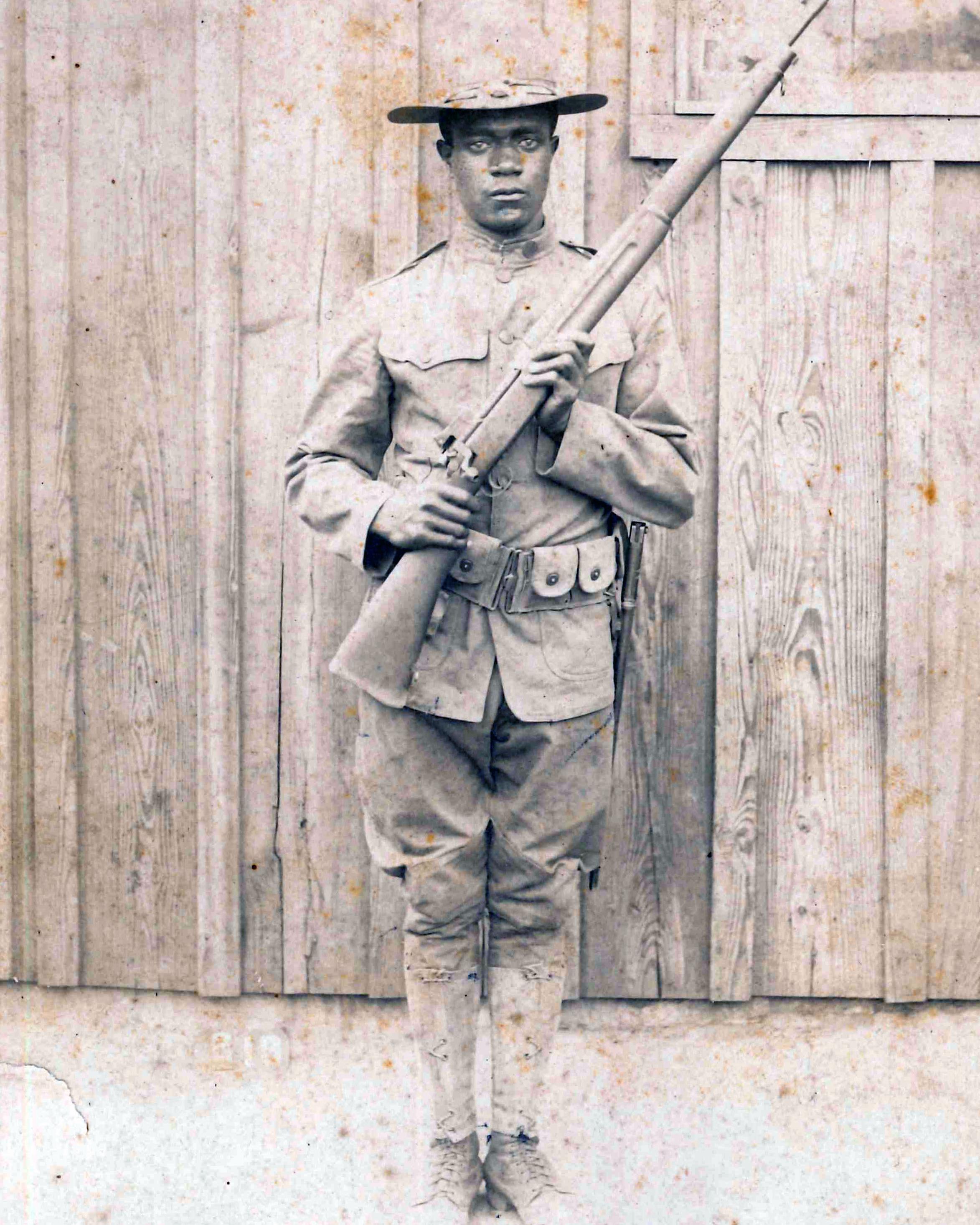 A young Black man stands wearing a U.S. World War I uniform and holding a rifle.