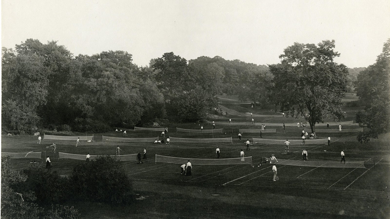 Black and white of many tennis courts and people on grassy area surrounded by trees