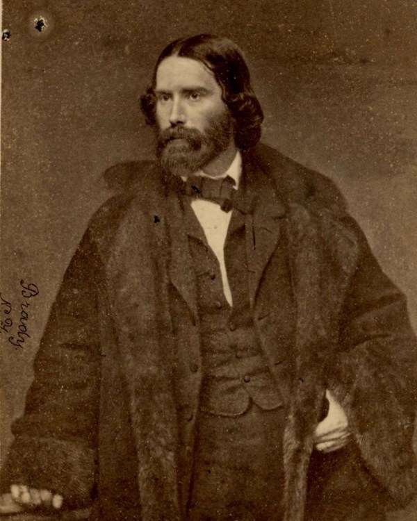 Black and white studio portrait of bearded man in suit and coat standing