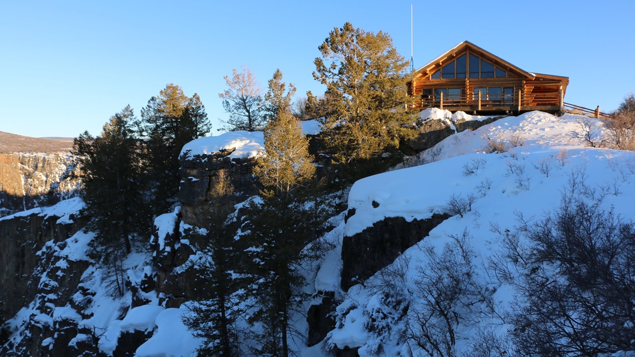 A log structure with large windows sits on the edge of a steep, snowy canyon wall.