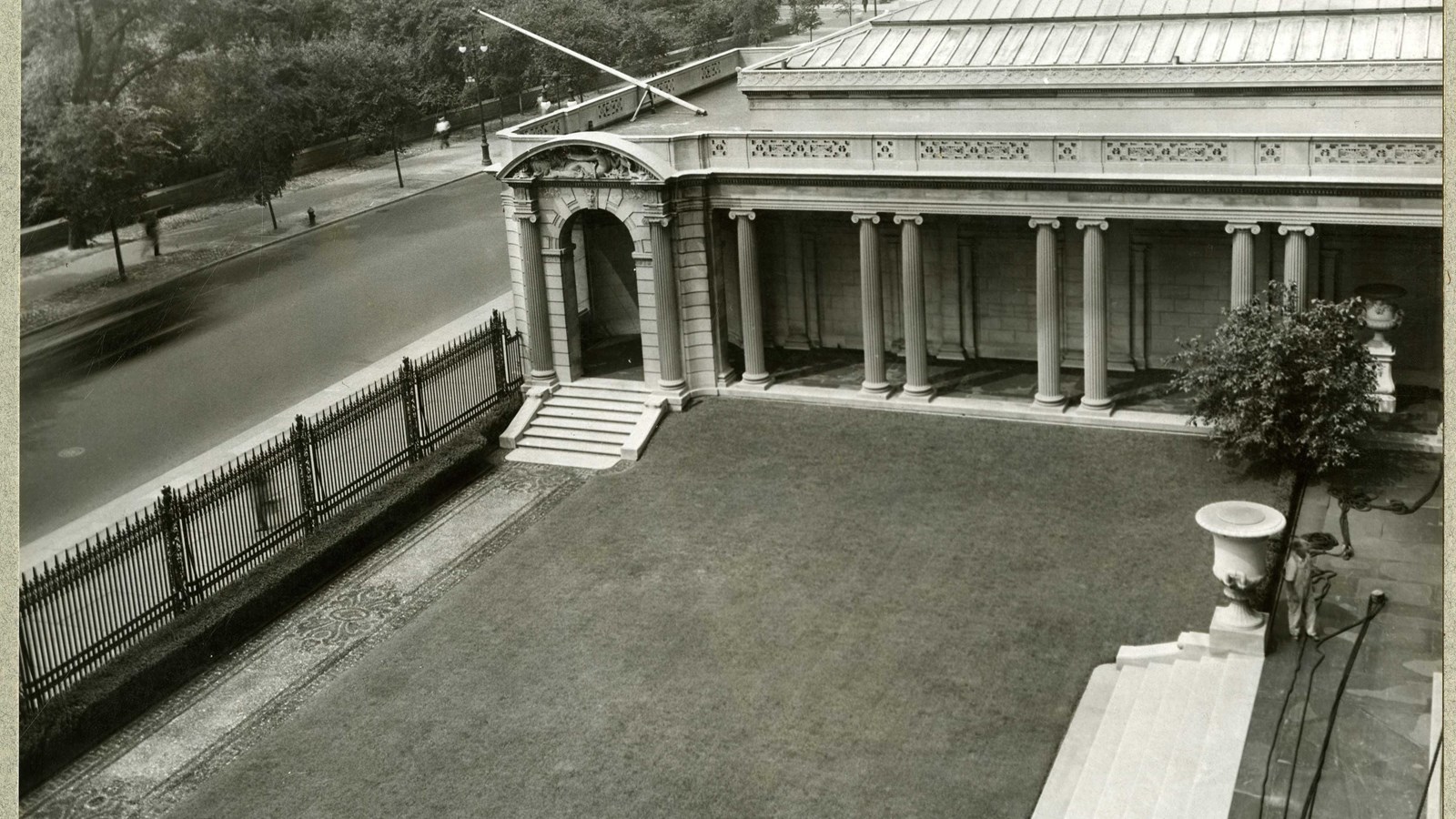 Black and white of flat grassy area enclosed by fence and building with road on other side