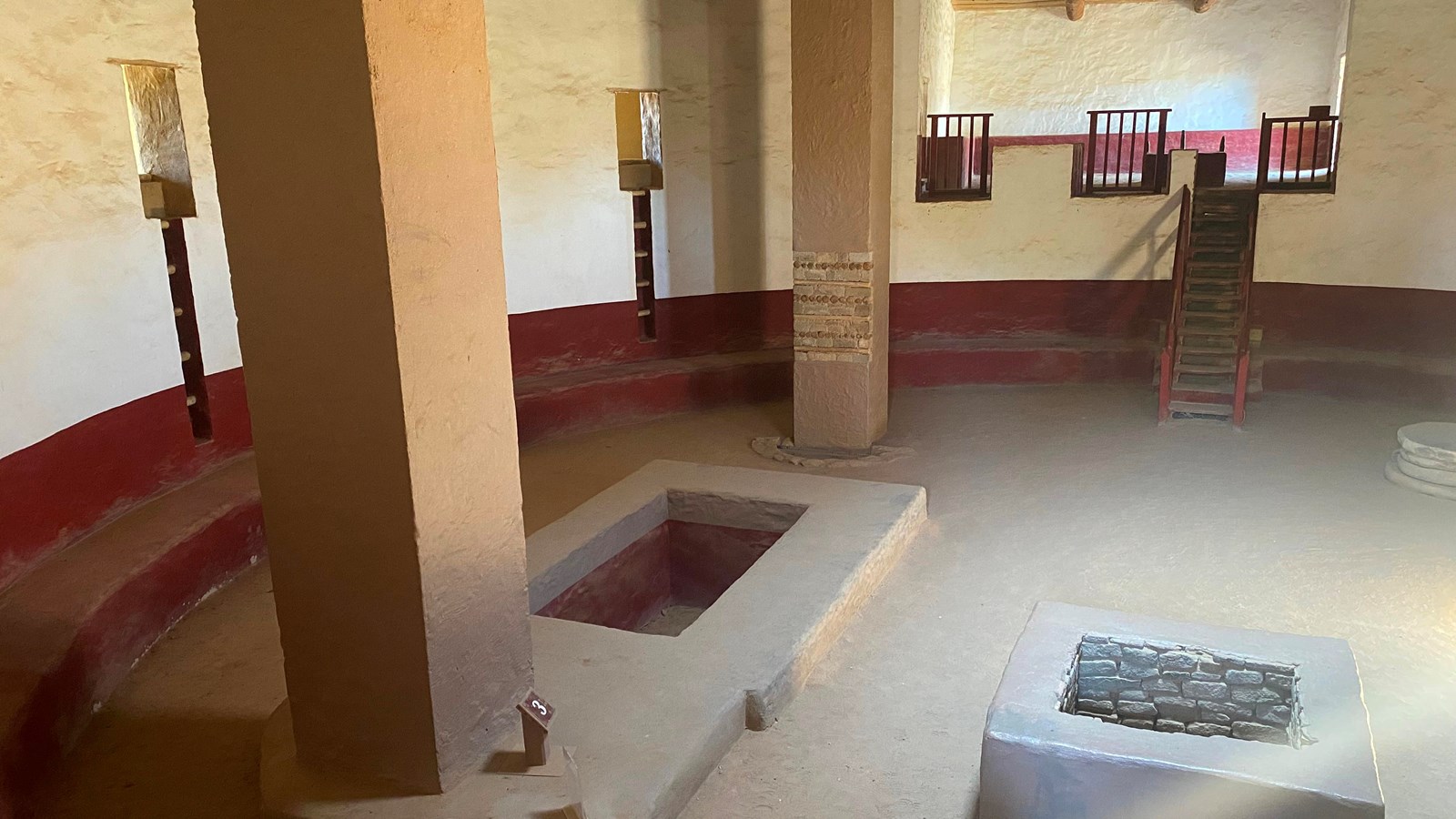 Half of the interior of a Great Kiva showing two columns, a bench, plastered walls, and a fire pit