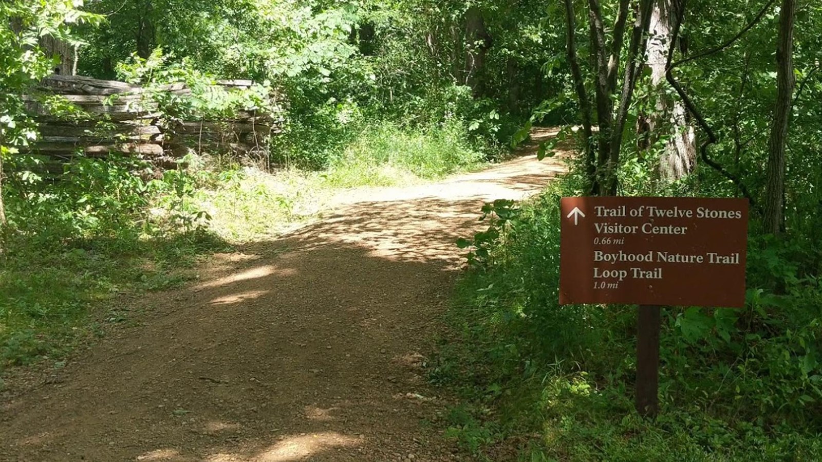 A sign reads Trail of Twelve Stones stands to the right of a forested trail.