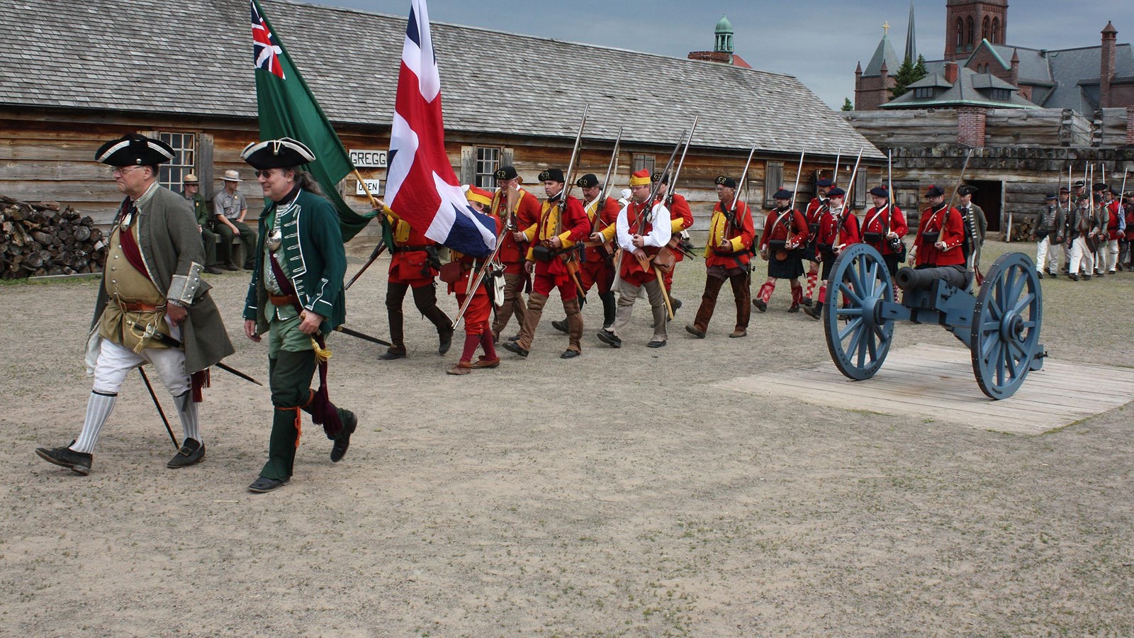 A group of soldiers wearing the same basic red uniform march in a line with a British flag in front.