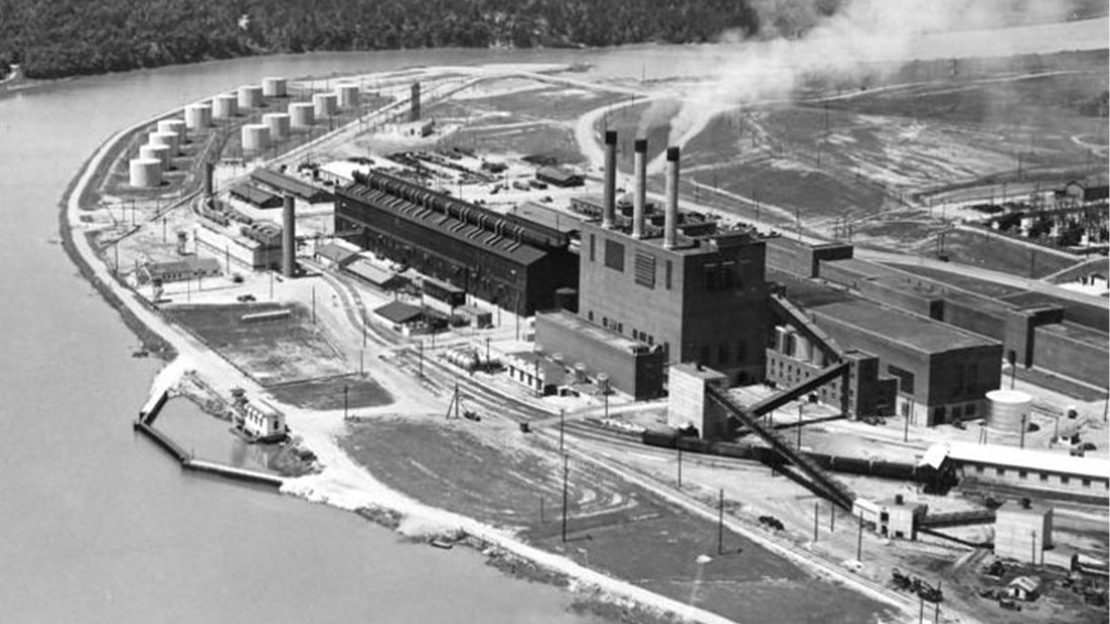 A large industrial complex with three smoking stacks rests on the bank of a winding river.