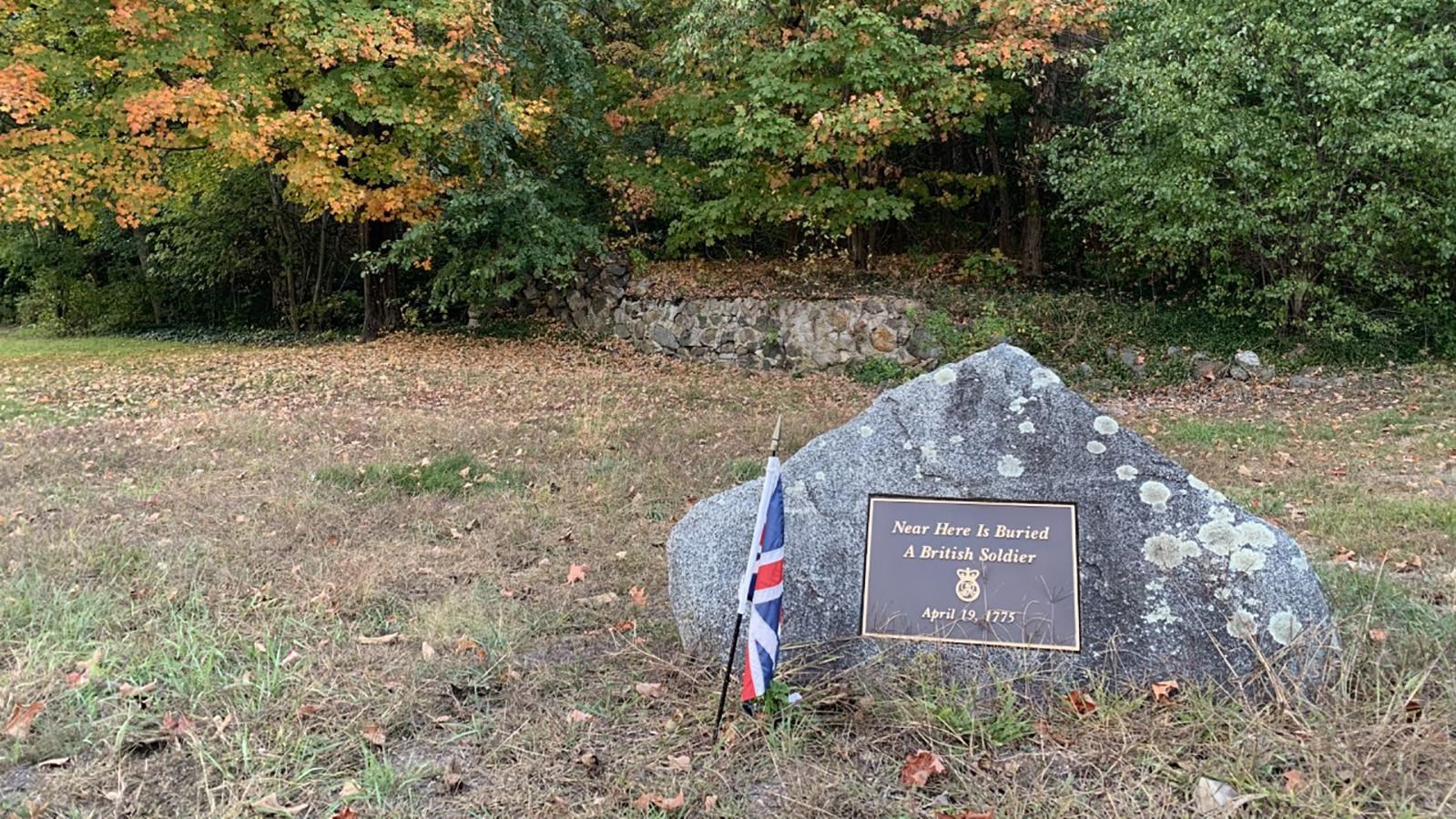 A stone grave marker for British soldiers sits before a tall stone fence in front of a tree lot.