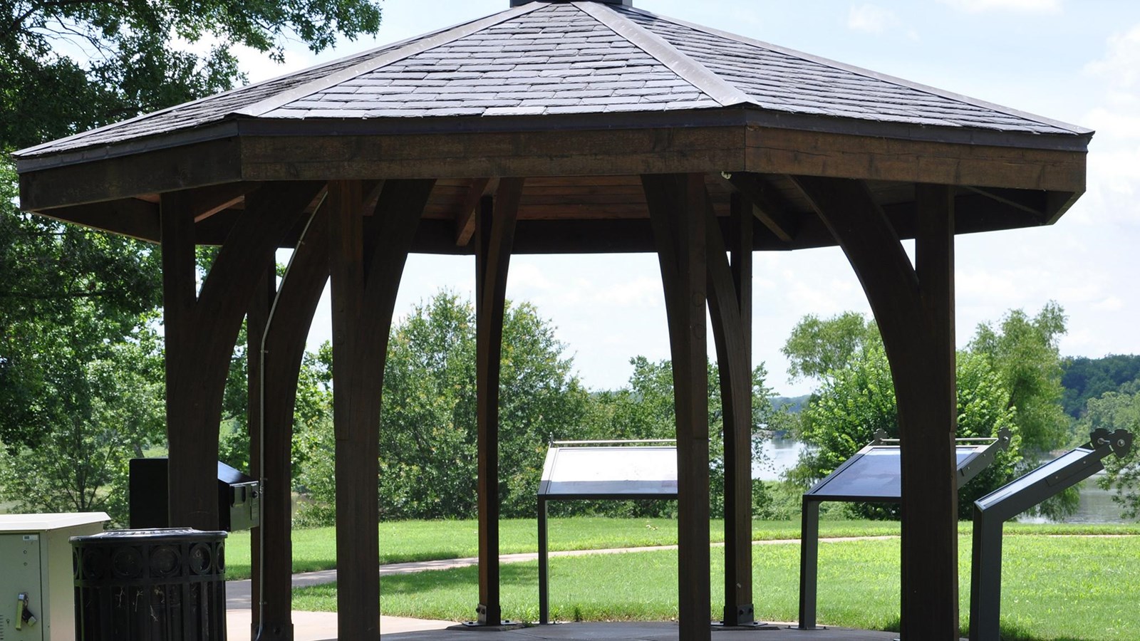 8 sided brown wooden gazebo with three waysides on a sunny day.
