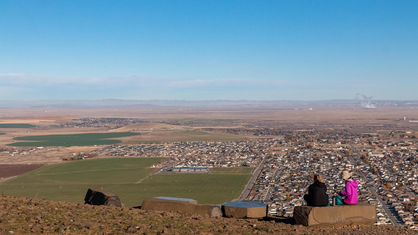 Two people sitting on a mountaintop overlooking a farmland and homes