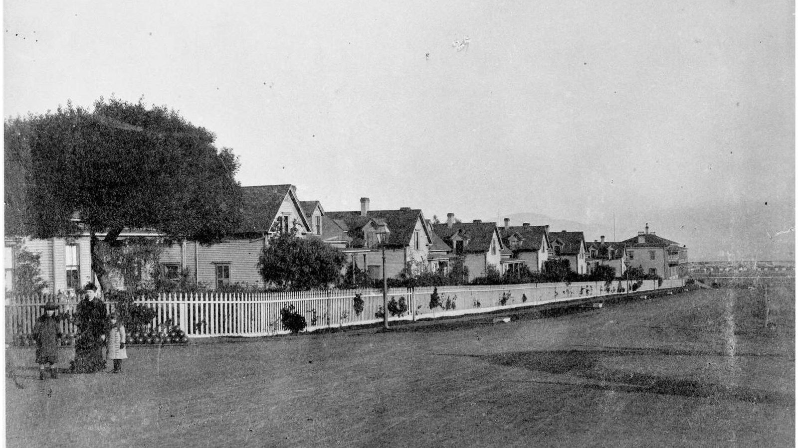 Black and white photo of one adult flanked by two children standing in front of a row of houses. 