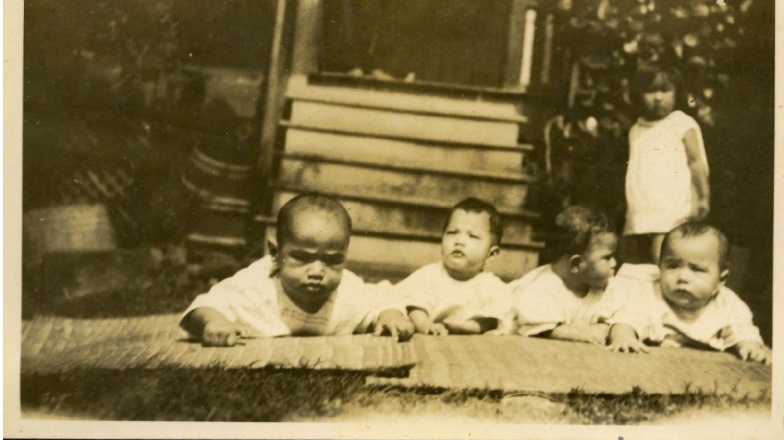 A black and white historic photo of four babies laying on the ground and a toddler behind.