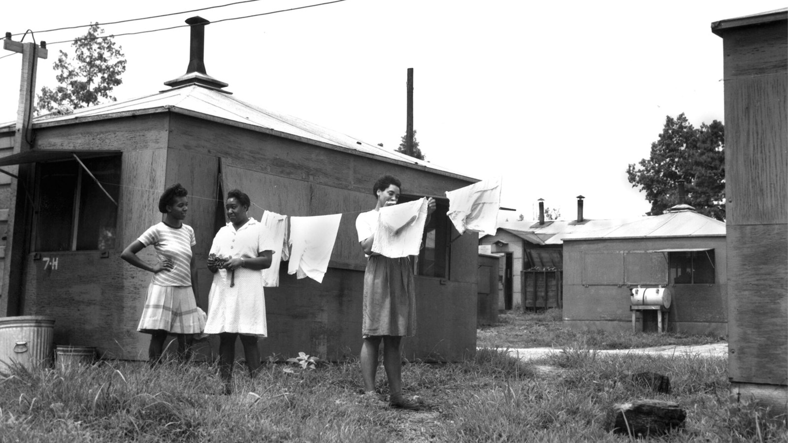 A black and white photo of 3 Black women with clothes on a clothesline between 2 small wooden shacks