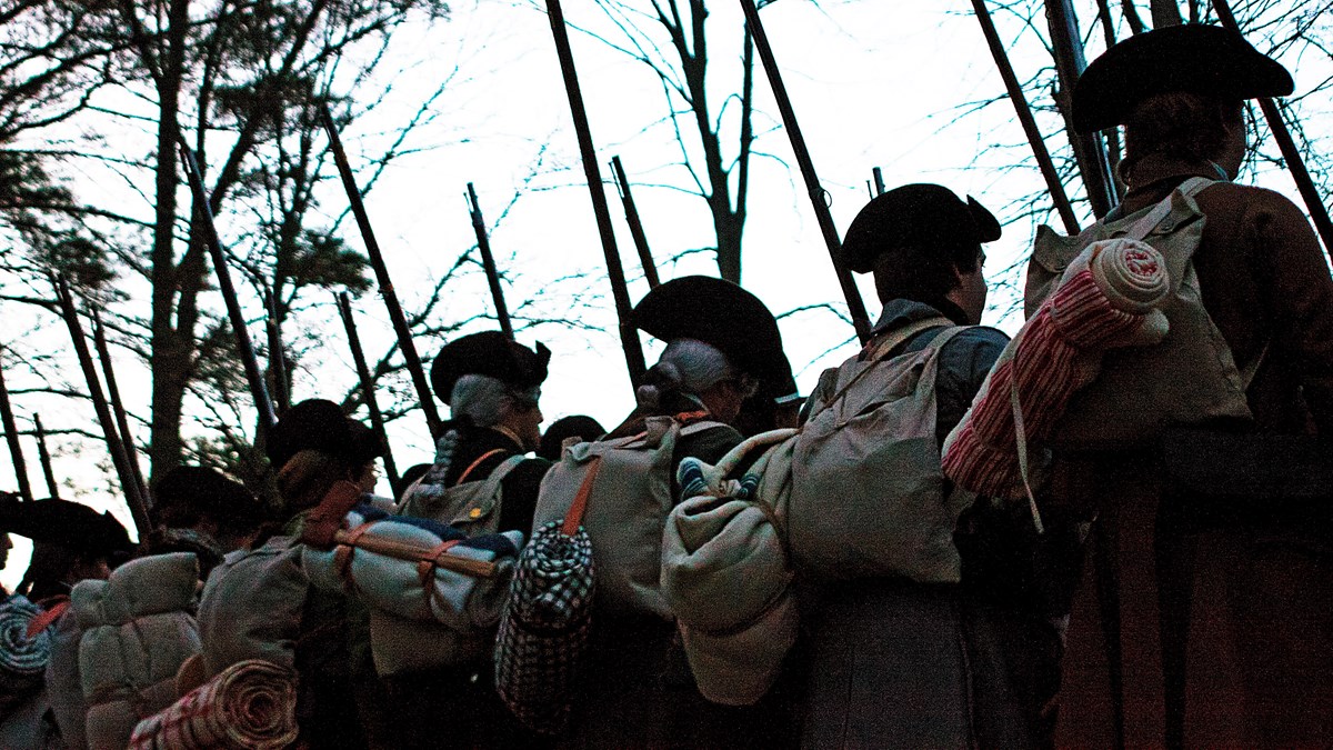 Several Colonial Revolutionary War militiamen viewed from behind stand in a line with muskets.
