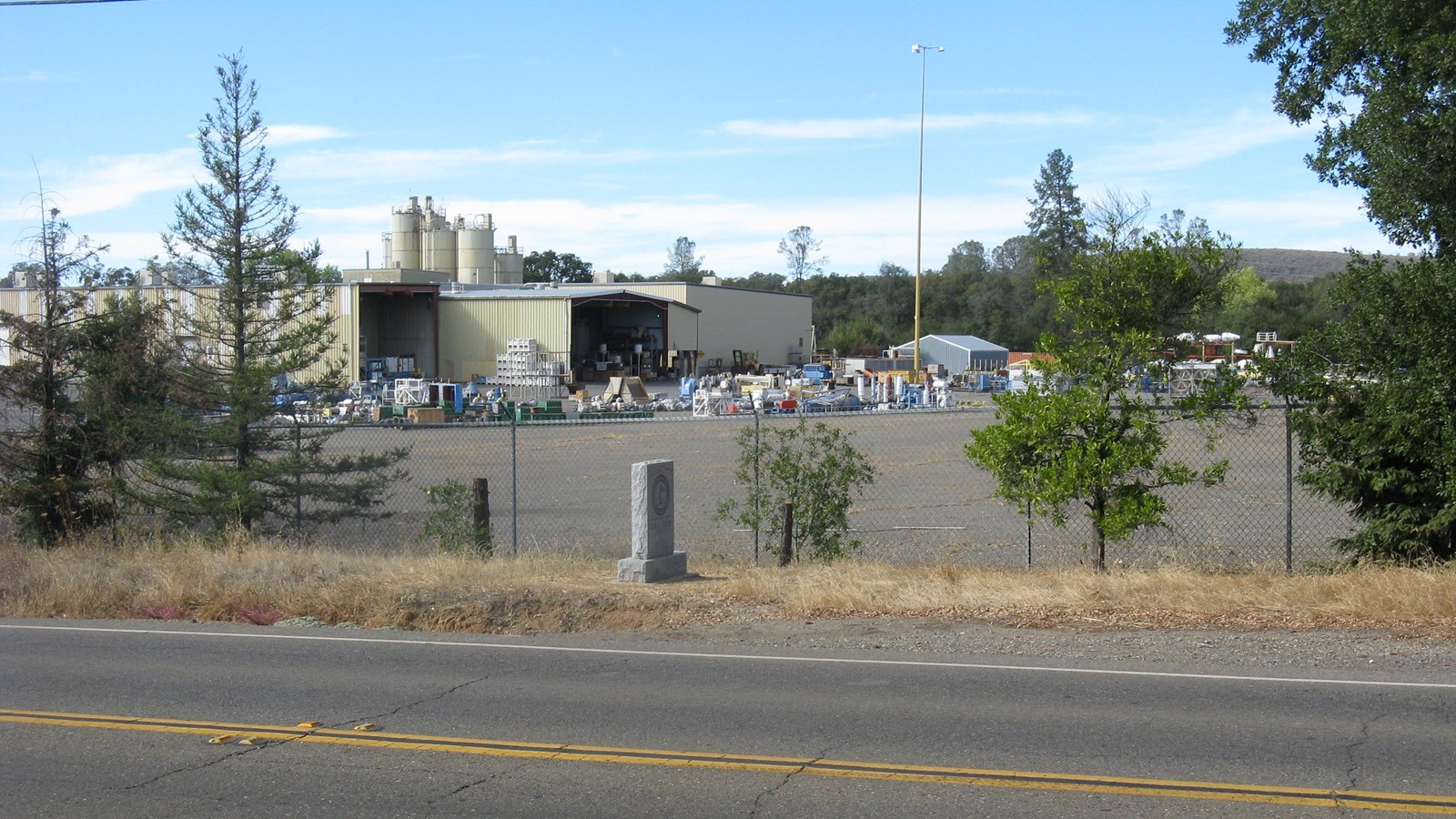 A gray, rectangular monument stands on a pedestal between the road pavement and a chain link fence. 