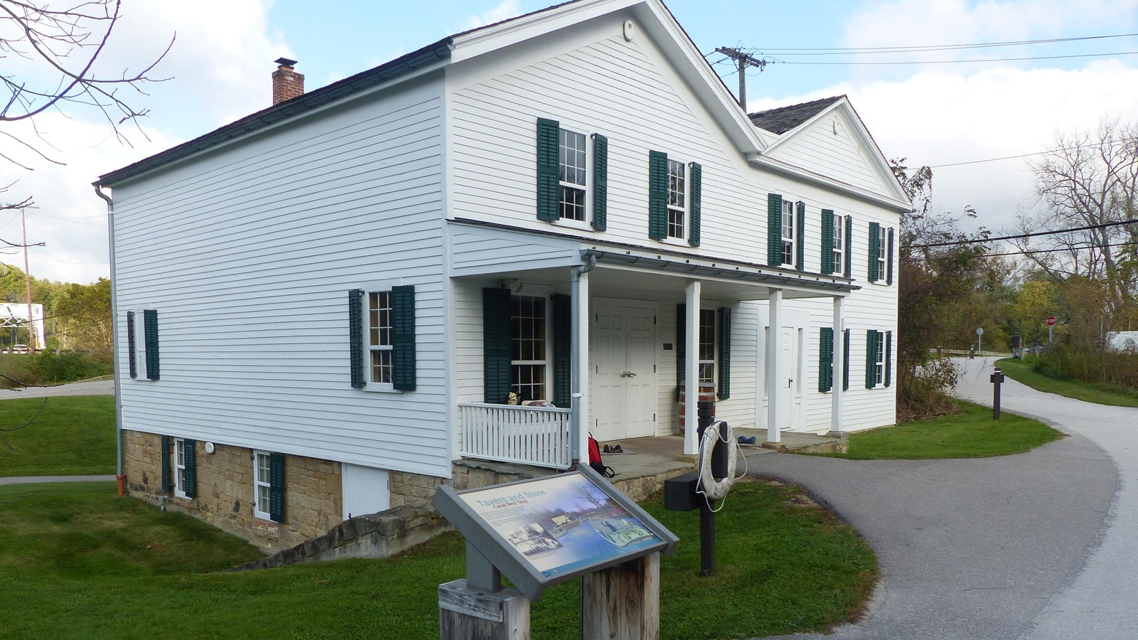 A graphic panel and life ring stand ouside a white historic building with a porch and an M-shaped