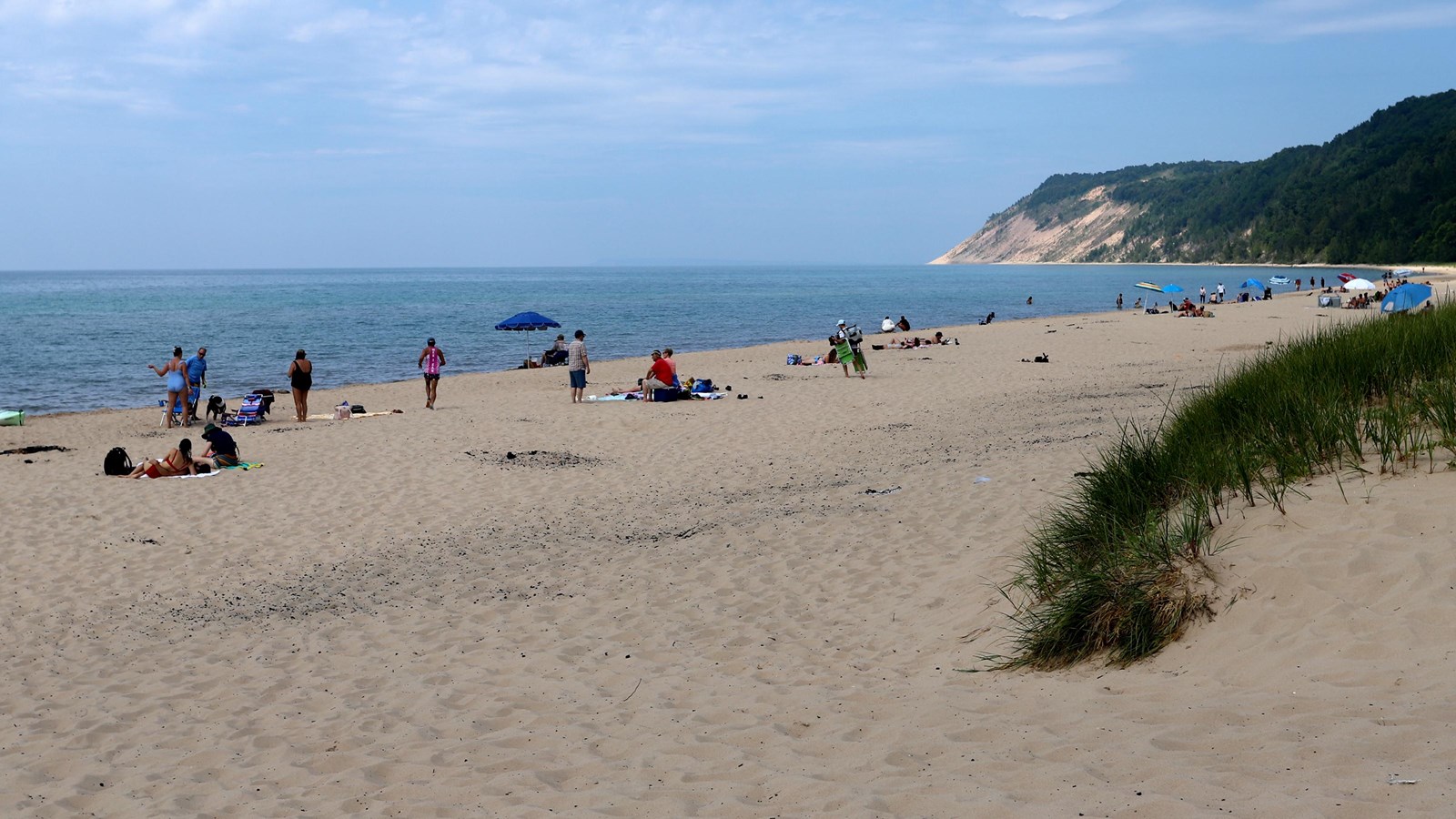 People in swimming gear sit on a sandy beach under a blue sky with dunes in the distance.