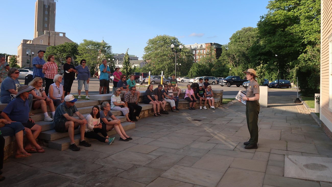 Ranger addressing large group of people sitting on stone steps in front of yellow building