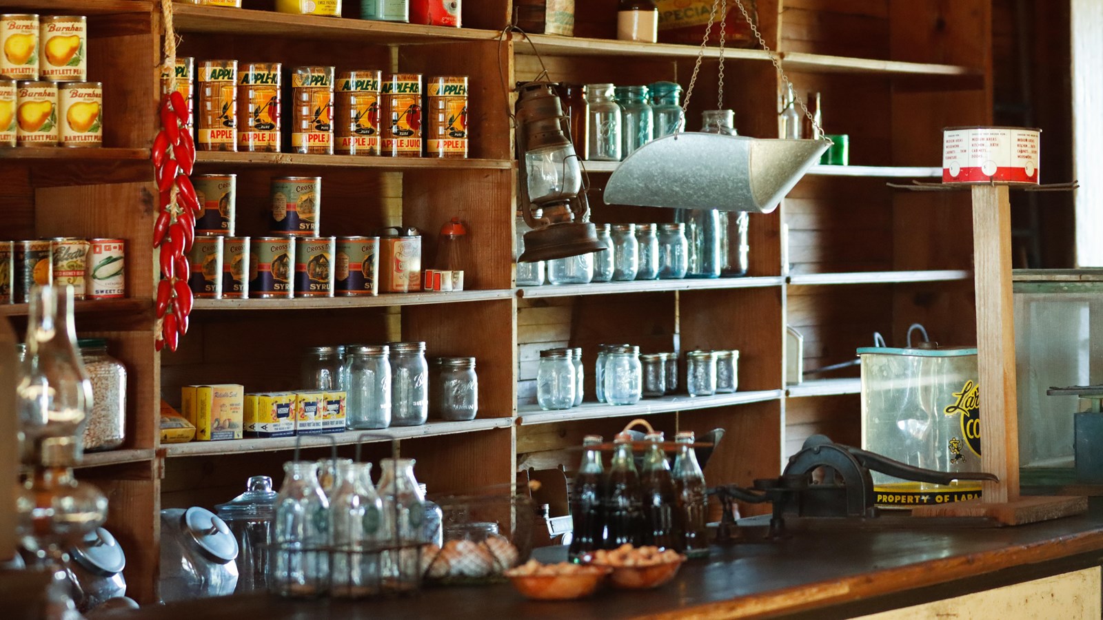 Interior of commissary. Canned goods, glass jars, and other food items sit on wood shelves. 