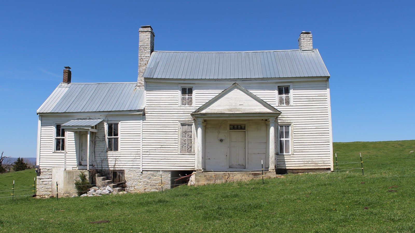 An 1800s farmhouse in a pasture has white clapboard siding and a grey roof.