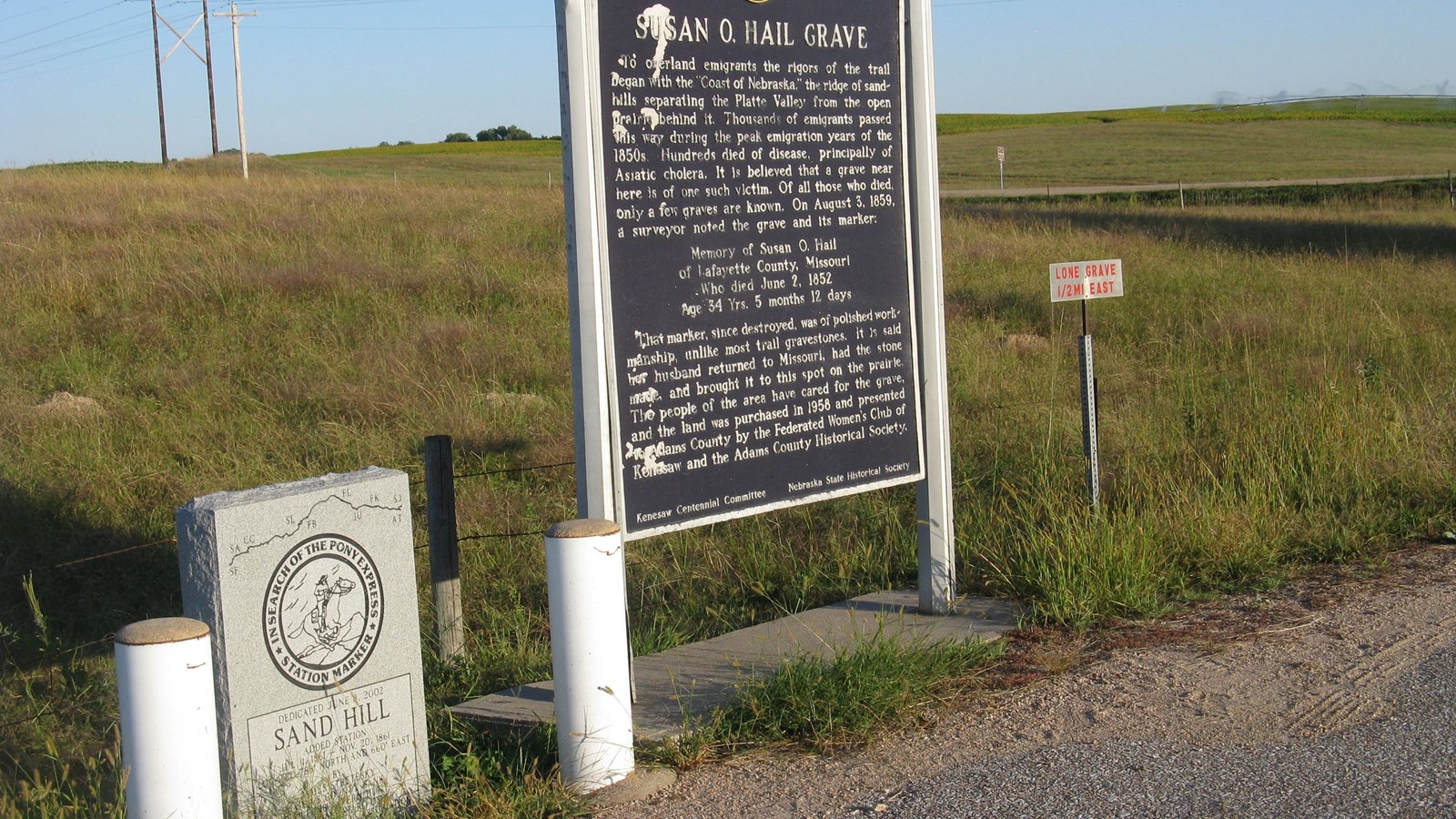 A large, brown sign panel with white text stands at the center. A smaller, granite monument to left.