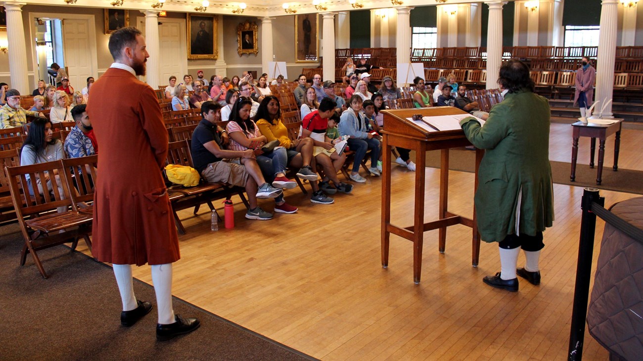 A large hall with a rangers, wearing colonial period clothing, speaking to the seated audience.