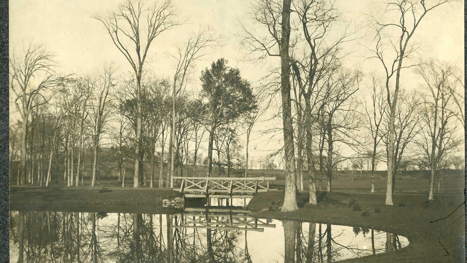 Black and white of body of water with bridge over one side and trees all around