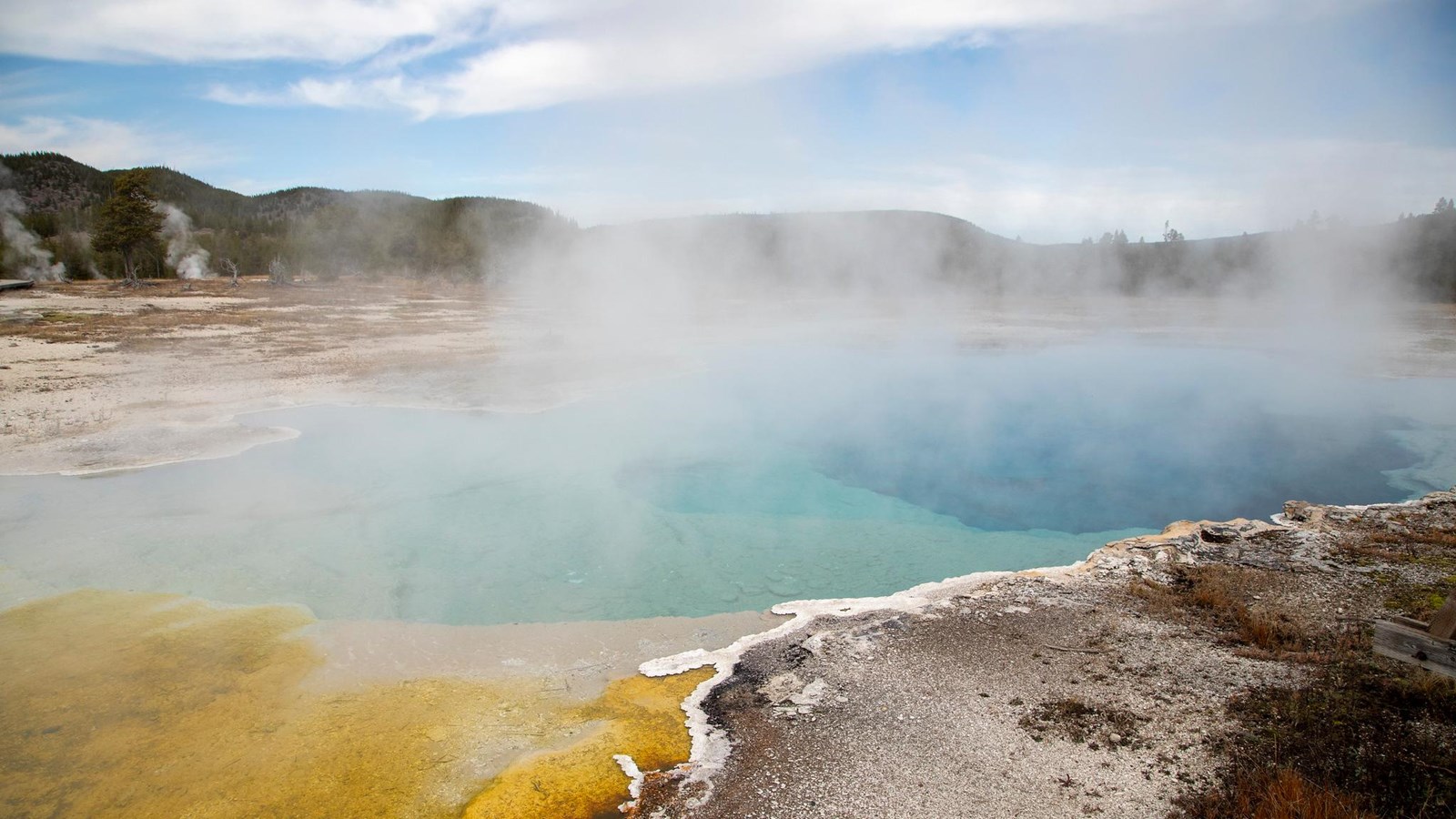 Steam rises off a turquoise blue hot spring with orange microbial mats along the edge.