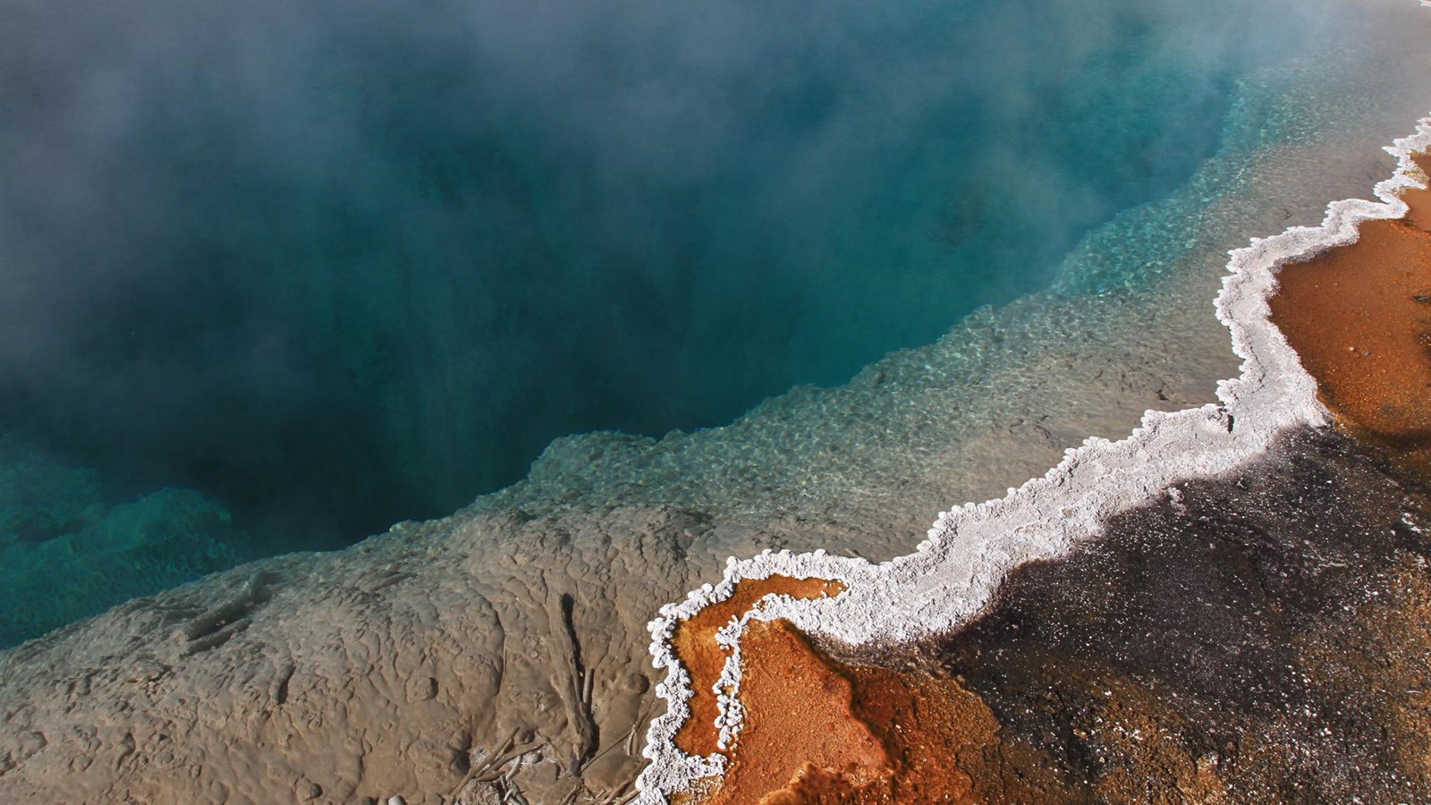 Steam rises off a turquoise blue hot spring with orange microbial mats along the edge.