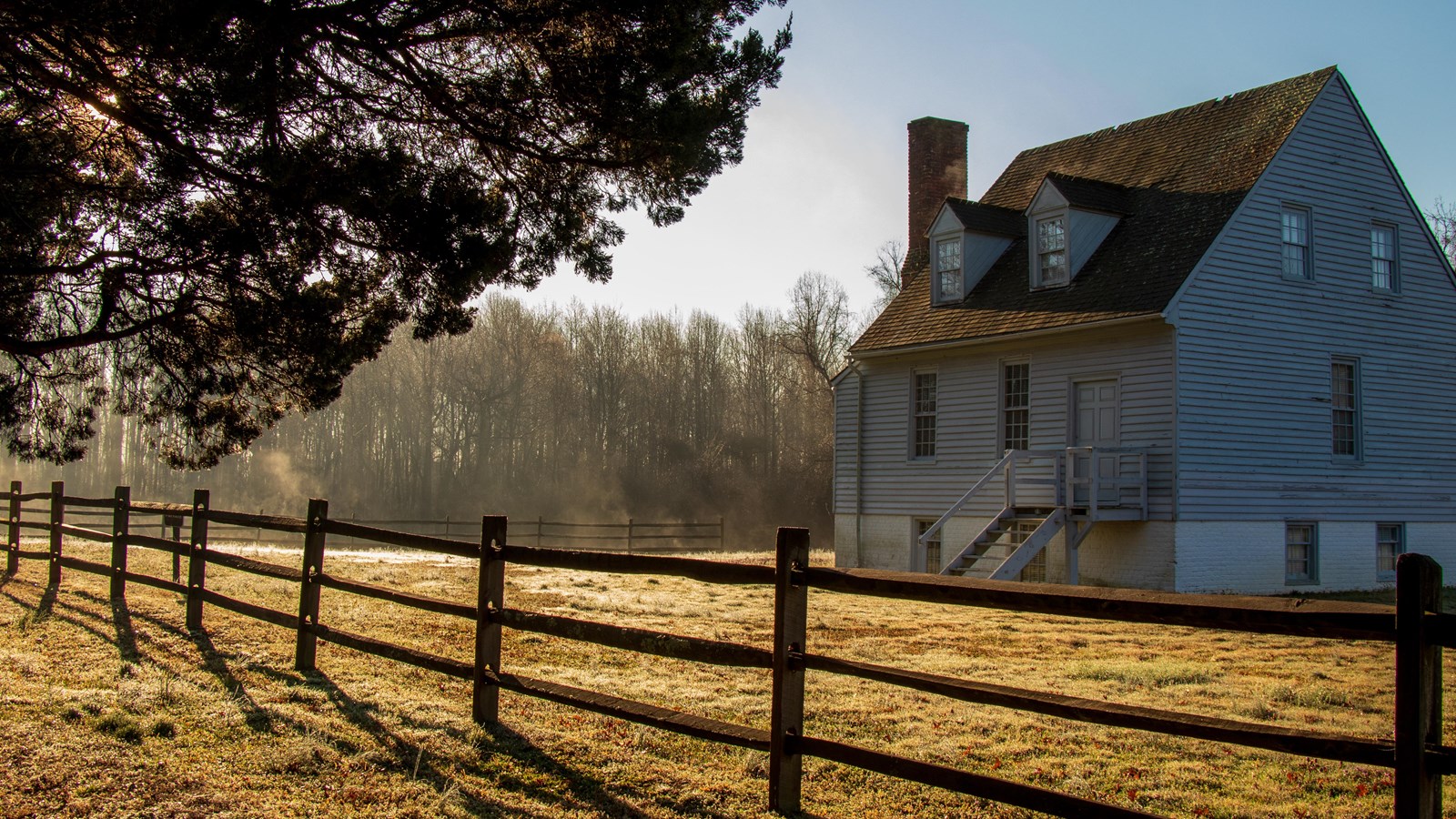 A small, wooden 19th-century house, painted gray, in a grassy field beside a wooden fence.